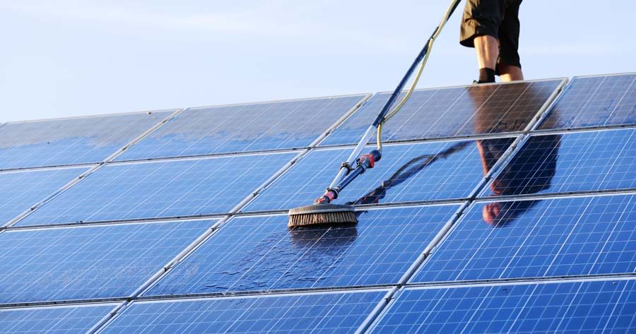 Technician soft-washing rooftop solar panels with a water-fed brush against a clear sky